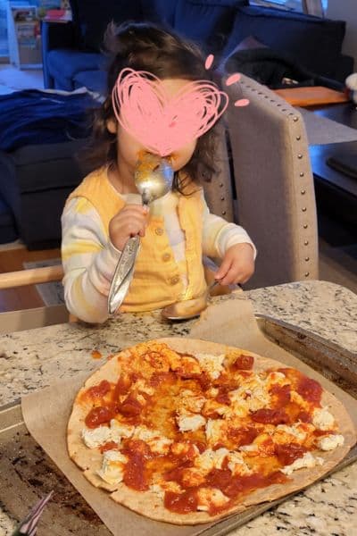 my toddler licking pizza sauce off of a spoon, in front of the pizza she made, as she kickstarts her cooking with toddlers phase